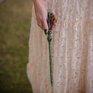 A close-up of a fairy wearing a pink lace dress and holding a dark green aventurine crystal wand outdoors.