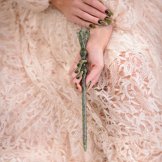 A close-up of a person in a pink lace dress holding a green aventurine crystal wand , with manicured green nails visible against the textured fabric.