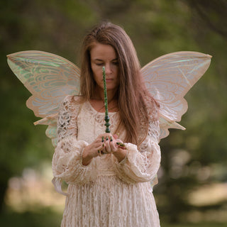 A fairy  in a long pink lace dress stands outdoors in a park-like setting with blurred green trees in the background. She has large translucent fairy wings attached to her back and is holding a green aventurine wand in her hands.