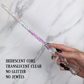 Handmade clear magic wand with  jewels and an iridescent core.
Held by a hand with black pointed finger nails on a white marble background. 