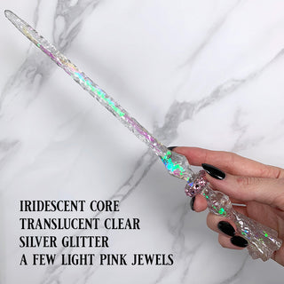 Handmade clear magic wand with  pink jewels, and iridescent core and glitter.

Held by a hand with black pointed finger nails on a white marble background. 