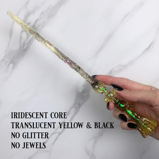 Handmade yellow and black magic wand with  an iridescent core.

Held by a hand with black pointed finger nails on a white marble background. 