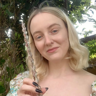 Grace from House of Wandcraft with blond hair and a floral dress holding a handmade wand in a leafy garden 