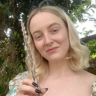 Grace from House of Wandcraft with blond hair and a floral dress holding a handmade wand in a leafy garden 