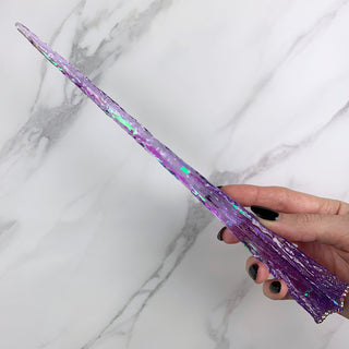 Handmade purple magic wand with  iridescent core and crystals. 
Held by a hand with black pointed finger nails on a white marble background. 