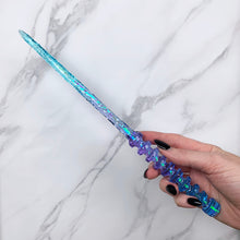 Load image into Gallery viewer, A stunning teal and purple celestial-inspired Orbit design magic wand featuring a delicate, translucent design with iridescent shimmering accents. The wand has a beautiful handle with alternating hoops with a narrow shaft that sparkles in light, showcasing its intricate craftsmanship. Perfect for fantasy collectors, this wand embodies elegance and enchantment, making it a prized addition to any magical assortment.
