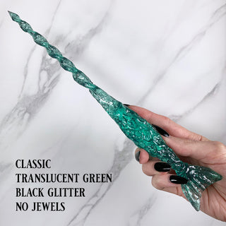 Handmade green magic wand with  black glitter. 
Held by a hand with black pointed finger nails on a white marble background. 