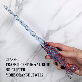 Handmade blue magic wand with  orange jewels. 
Held by a hand with black pointed finger nails on a white marble background. 