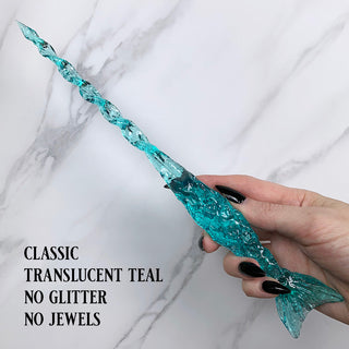 Handmade teal magic wand .

Held by a hand with black pointed finger nails on a white marble background. 