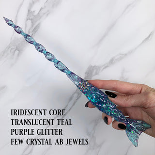 Handmade teal magic wand with  purple glitter and jewels. 
Held by a hand with black pointed finger nails on a white marble background. 