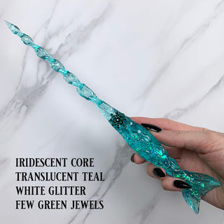 Handmade teal magic wand with  white glitter and green jewels. 
Held by a hand with black pointed finger nails on a white marble background. 