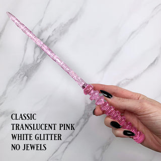 Handmade pink magic wand with  white glitter.
 
Held by a hand with black pointed finger nails on a white marble background. 