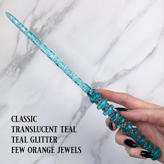 Handmade teal magic wand with  teal glitter and orange jewels. 

Held by a hand with black pointed finger nails on a white marble background. 