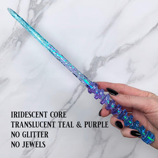 Handmade teal and purple magic wand with iridescent core.
 
Held by a hand with black pointed finger nails on a white marble background. 