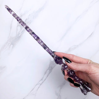 A unique handmade magic wand made with real purple amethyst crystal chips, in the Flora Design. 
On a white marbled background being held by a hand with black pointed nails. 