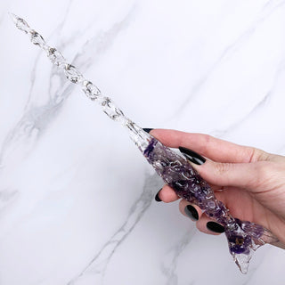A unique handmade magic wand made with real purple amethyst crystal chips, in the Mertail  Design. 
On a white marbled background being held by a hand with black pointed nails. 
