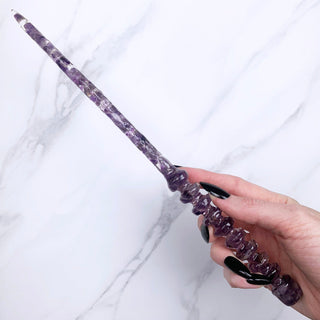 A unique handmade magic wand made with real purple amethyst crystal chips, in the Orbit  Design. 

On a white marbled background being held by a hand with black pointed nails. 