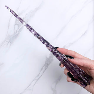 A unique handmade magic wand made with real purple amethyst crystal chips, in the Stat  Design. 

On a white marbled background being held by a hand with black pointed nails. 
