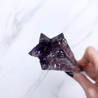 A unique handmade magic wand made with real purple amethyst crystal chips, in the Star  Design. 

The detail of the end of the wand is a five pointed star and is on a white marbled background being held by a hand with black pointed nails. 