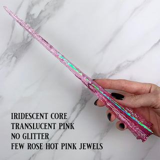 Handmade pink magic wand with  iridescent core and pink jewels. 
Held by a hand with black pointed finger nails on a white marble background. 