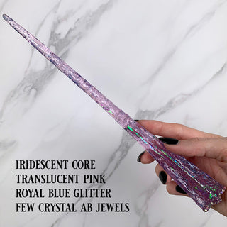 Handmade pink magic wand with  an iridescent core, blue glitter and AB jewels. 
Held by a hand with black pointed finger nails on a white marble background. 