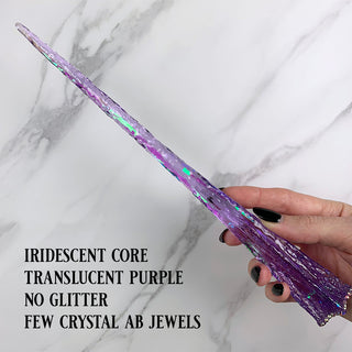 Handmade purple magic wand with  iridescent core and AB jewels.
 
Held by a hand with black pointed finger nails on a white marble background. 