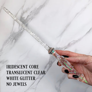 Handmade clear magic wand with  white glitter and an iridescent core. 
Held by a hand with black pointed finger nails on a white marble background. 