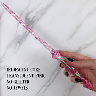 Handmade pink magic wand with  an iridescent core. 
Held by a hand with black pointed finger nails on a white marble background. 