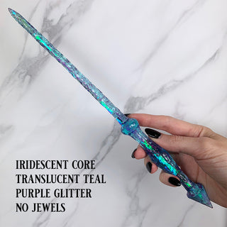 Handmade teal magic wand with  iridescent core and purple glitter.
 
Held by a hand with black pointed finger nails on a white marble background. 