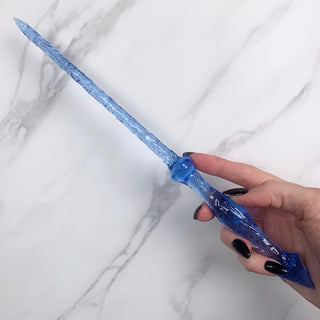 Handmade blue magic wand. 
Held by a hand with black pointed finger nails on a white marble background. 