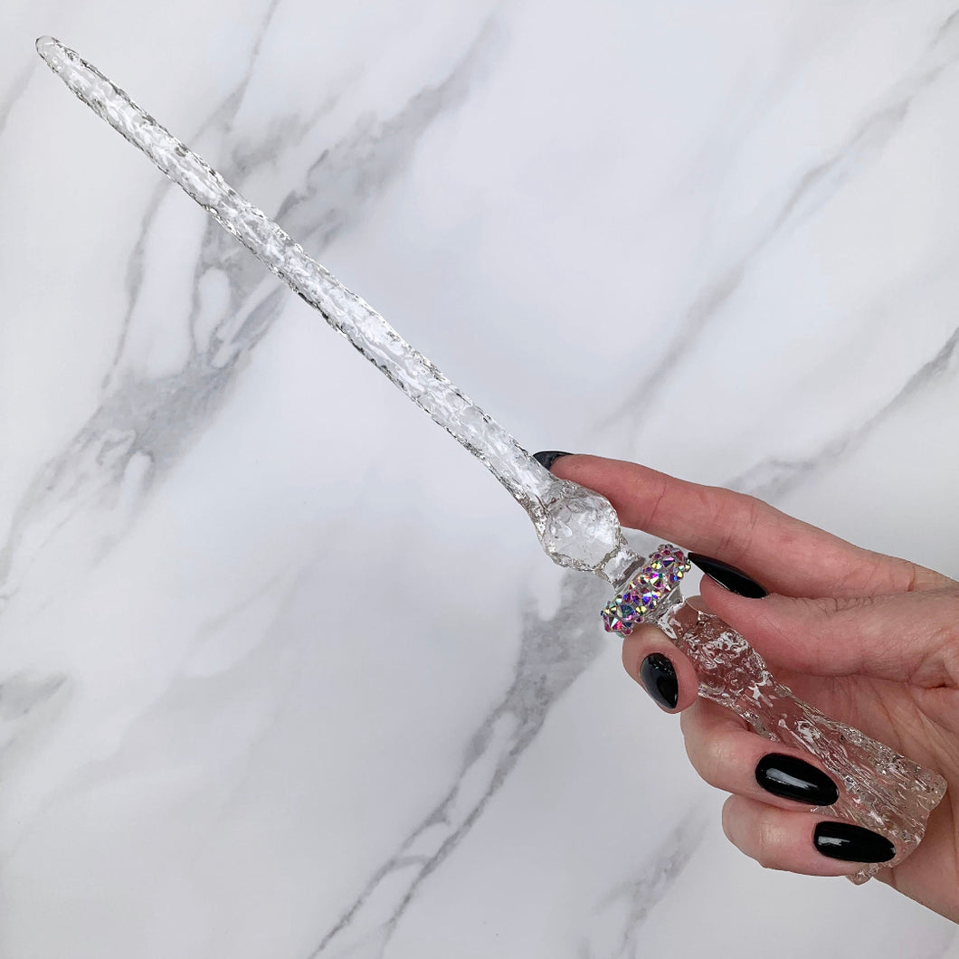 A stunning clear Flora magic wand featuring a delicate, translucent design with iridescent shimmering accents. The wand has a beautiful flower shaped base with a narrow shaft that sparkles in light, showcasing its intricate craftsmanship. Perfect for fantasy collectors, this wand embodies elegance and enchantment, making it a prized addition to any magical assortment.