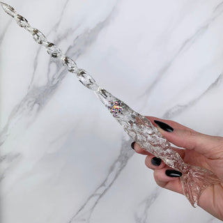 Handmade clear magic wand with  jewels. 
Held by a hand with black pointed finger nails on a white marble background. 