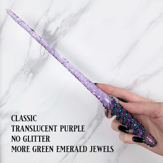 Handmade purple magic wand with  green jewels. 
Held by a hand with black pointed finger nails on a white marble background. 