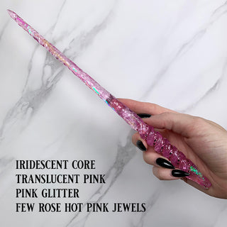 Handmade pink  magic wand with  an iridescent core, pink glitter and pink jewels. 

Held by a hand with black pointed finger nails on a white marble background. 