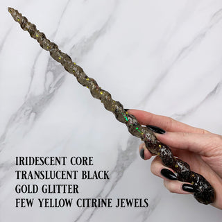 Handmade black magic wand with  iridescent core, gold glitter and yellow jewels. 

Held by a hand with black pointed finger nails on a white marble background. 