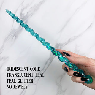 Handmade teal magic wand with  iridescent core and teal glitter. 

Held by a hand with black pointed finger nails on a white marble background. 