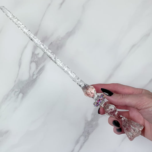 A stunning clear Flora magic wand featuring a delicate, translucent design with iridescent shimmering accents. The wand has a beautiful flower shaped base with a narrow shaft that sparkles in light, showcasing its intricate craftsmanship. Perfect for fantasy collectors, this wand embodies elegance and enchantment, making it a prized addition to any magical assortment.