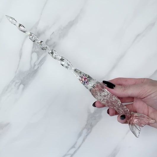 A stunning clear Mermaid inspired magic wand featuring a delicate, translucent design with iridescent shimmering accents. The wand has a beautiful fishtail shaped base with a swirling shaft that sparkles in light, showcasing its intricate craftsmanship. Perfect for fantasy collectors, this wand embodies elegance and enchantment, making it a prized addition to any magical assortment.
