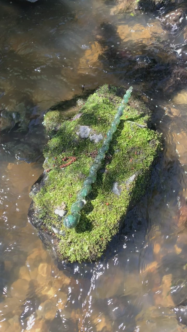 A unique handmade magic wand made with real green aventurine crystal chips, in the unicorn Design. 
The background is a moss covered stone  a whimsical river. 